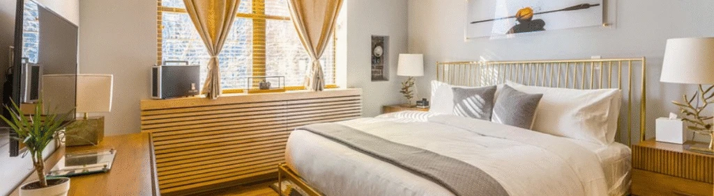 Modern bedroom with a large bed, two lamps, sunlight through curtained windows, and neutral decor.