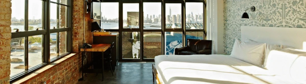 Sunny loft bedroom with brick walls, large windows, city skyline view, and modern decor.