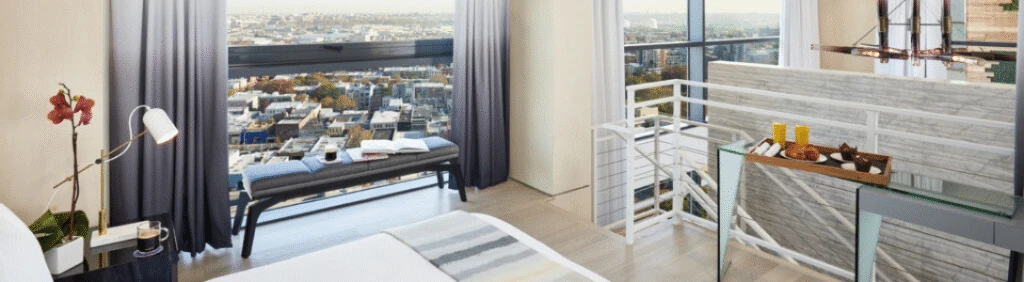 Modern hotel room with a large window, city view, a bench, and breakfast setup on a small table.