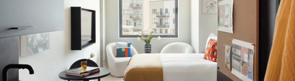 Modern, cozy bedroom with a double bed, colorful pillows, round chair, and large window letting in daylight.