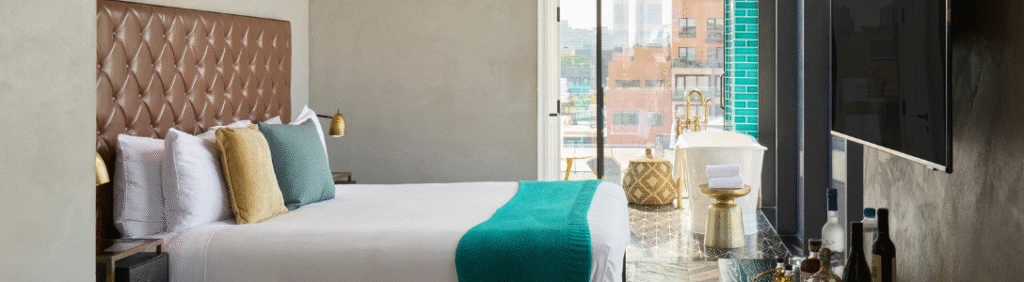 Modern hotel room with a large bed, teal accents, and a freestanding bathtub by a window with city views.