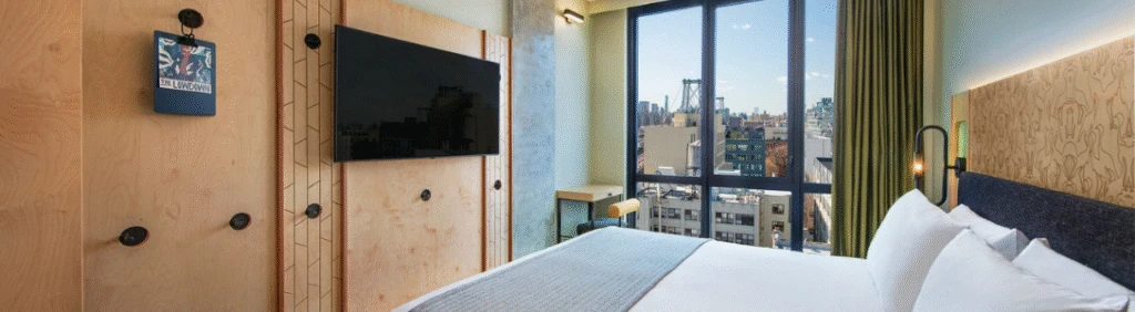 Modern hotel room with a large bed, wall-mounted TV, and a window offering a city skyline view.