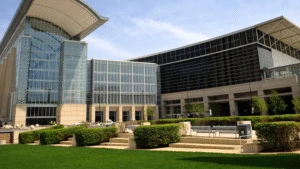 Modern glass and steel convention center with landscaped green lawn and trimmed bushes in the foreground.
