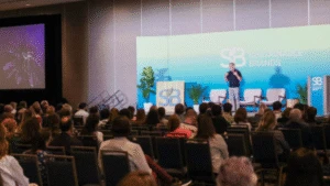 A speaker presents on stage to an audience at a Sustainable Brands conference.