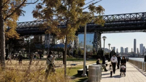 People walk and relax by the river under a large bridge on a sunny day with city buildings in the background.